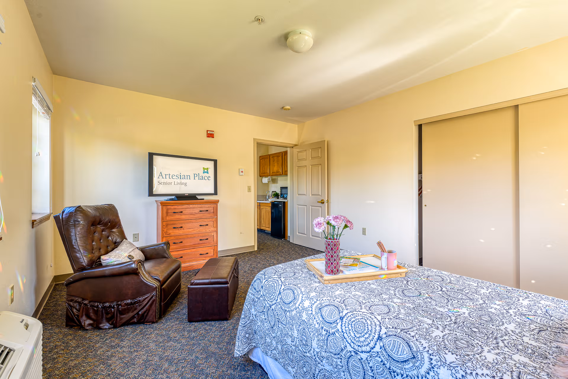 A cozy senior living room featuring a patterned bedspread on a bed, a wooden dresser with a TV displaying 'Artesian Place Senior Living,' a brown leather armchair with a pillow, a small ottoman, and a tray with flowers and a cup on the bed. The room has beige walls, a window with blinds, a closet with sliding doors, and an open door leading to a kitchen area.