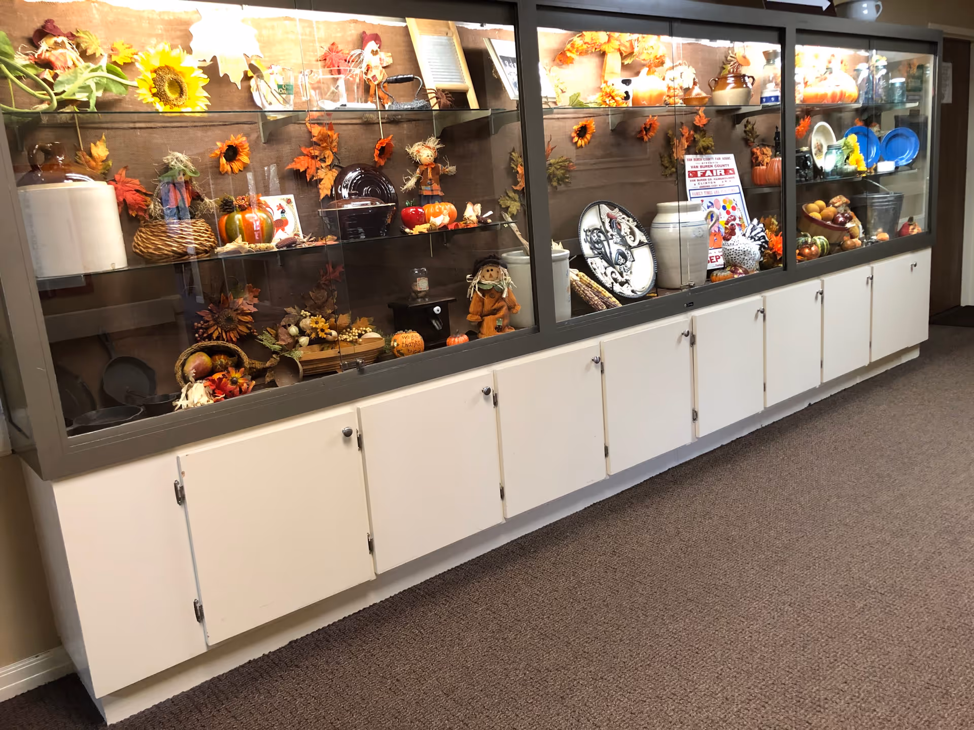A long display cabinet with glass doors showcasing various autumn-themed decorations including sunflowers, pumpkins, scarecrows, ceramic dishes, and other fall decor items. The cabinet has white lower storage doors and is situated on a carpeted floor.