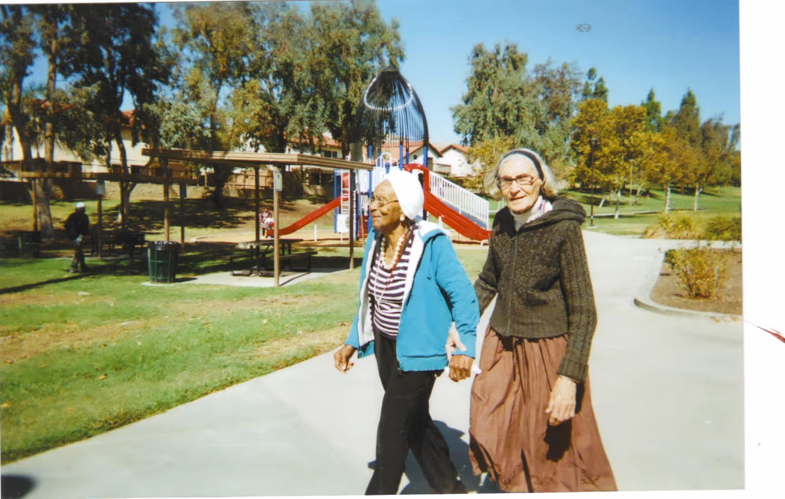 Two elderly women walking hand in hand on a paved path in a park-like outdoor area with green grass, trees, and a playground with a red slide in the background under a clear blue sky.