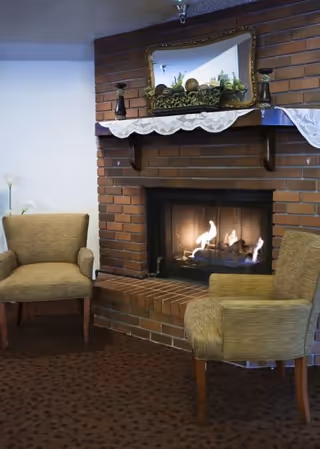 A cozy indoor seating area with two beige armchairs positioned around a lit brick fireplace. Above the fireplace is a mantle decorated with a lace runner, two candle holders, and a large mirror reflecting some greenery and decorative items.