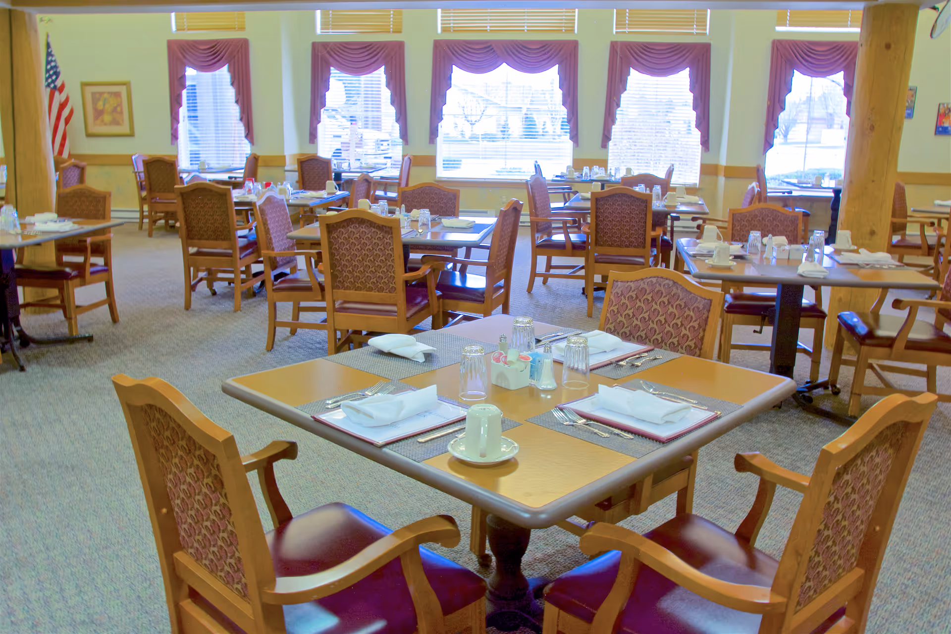 A dining room in a senior living facility with multiple wooden tables and chairs arranged neatly. Each table is set with placemats, napkins, cups, glasses, and silverware. Large windows with maroon valances allow natural light to fill the room. An American flag and framed artwork are visible on the walls.
