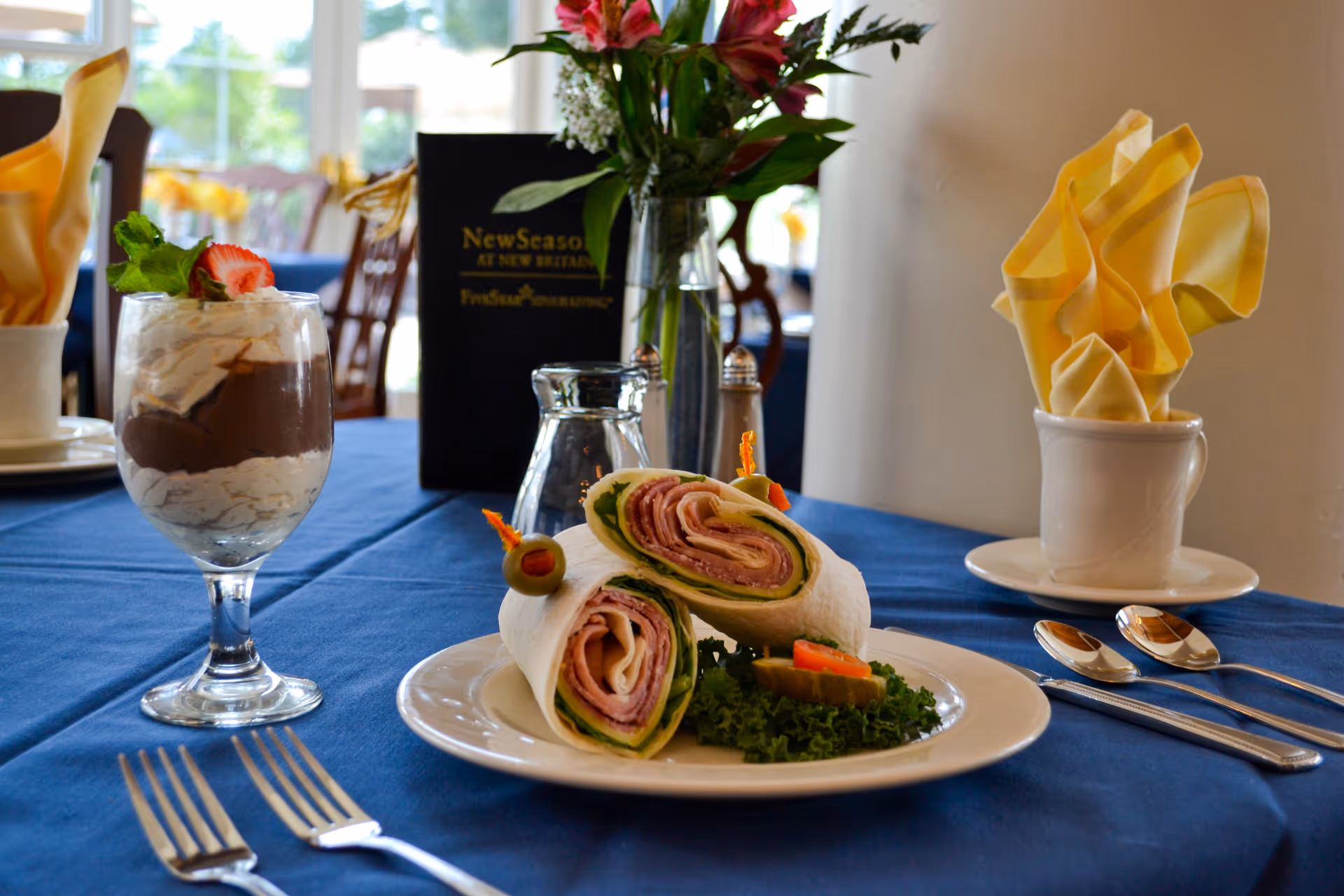 A dining table set with a plate of two wrap sandwiches garnished with olives and lettuce, a glass dessert with chocolate and whipped cream topped with a strawberry slice, silverware, and a yellow folded napkin in a white cup. In the background, there is a menu card with the text 'New Seasons At New Britain' and a vase with flowers.
