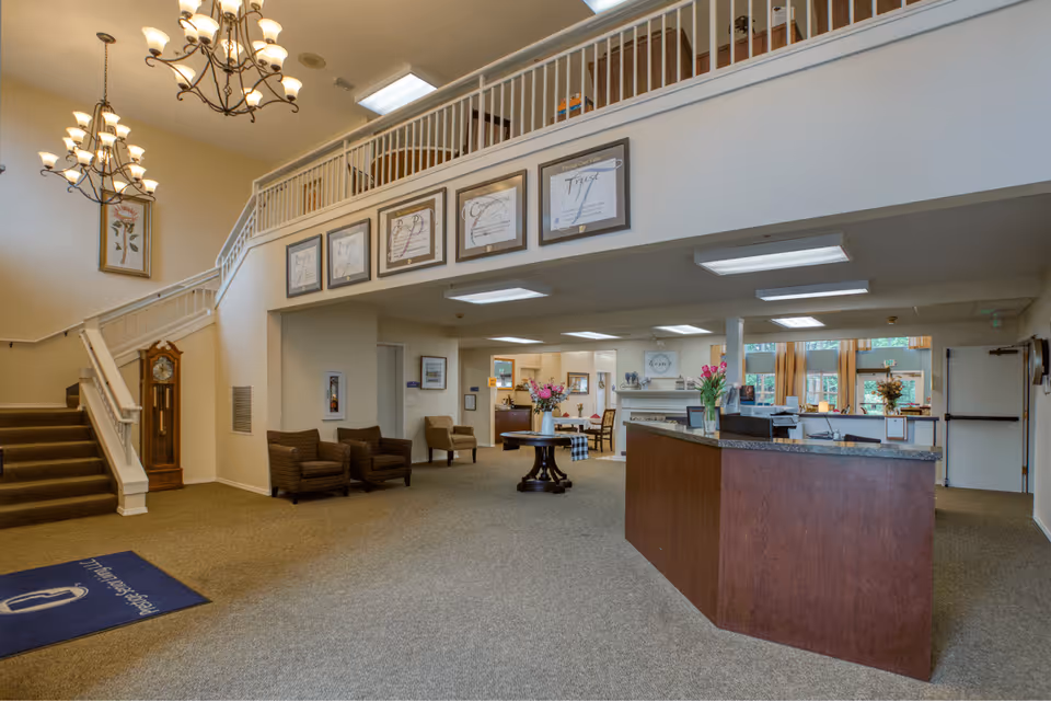 Interior view of a senior living facility lobby with a reception desk on the right, a staircase with white railings on the left, several chairs along the walls, framed artwork and inspirational quotes hanging above the reception area, and chandeliers hanging from the ceiling.