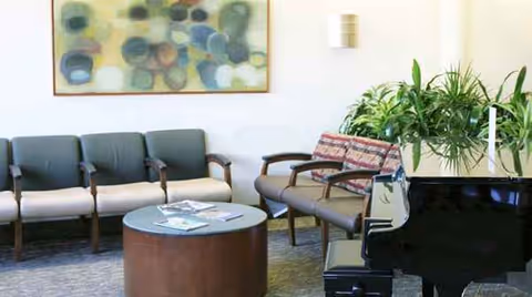 Lounge area with row of chairs, round coffee table, piano, plant, and abstract art.