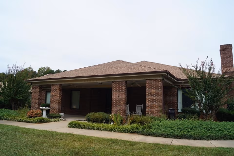 Exterior view of a single-story brick building with a covered porch supported by brick columns. The porch has two white rocking chairs and is surrounded by well-maintained shrubs and greenery. A concrete walkway leads up to the porch, and the sky is overcast.
