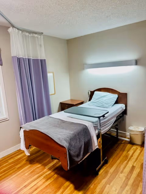 A small, simple patient room with a single hospital bed made with white sheets, a gray blanket, and a pillow. The bed has an overbed table positioned on it. There is a wooden nightstand next to the bed, a trash can on the floor, and a window with purple and white curtains. The room has wooden flooring and a wall-mounted light above the bed.