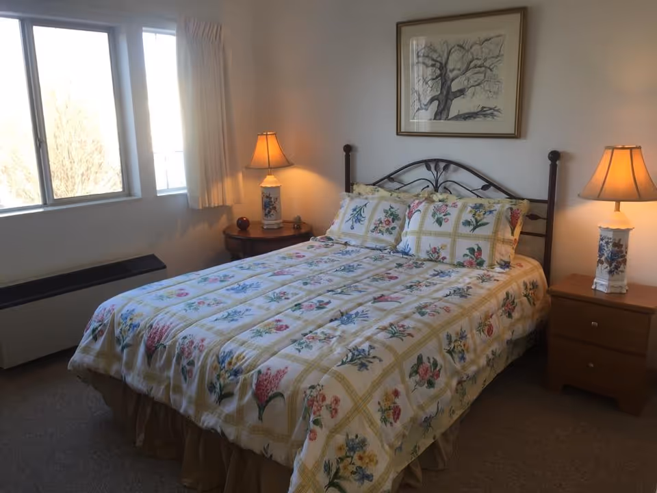 A cozy bedroom with a floral-patterned bedspread on a double bed. The bed has a metal headboard and is flanked by two wooden nightstands, each with a decorative lamp. A framed picture of a tree hangs on the wall above the bed. There is a window with cream-colored curtains letting in natural light.