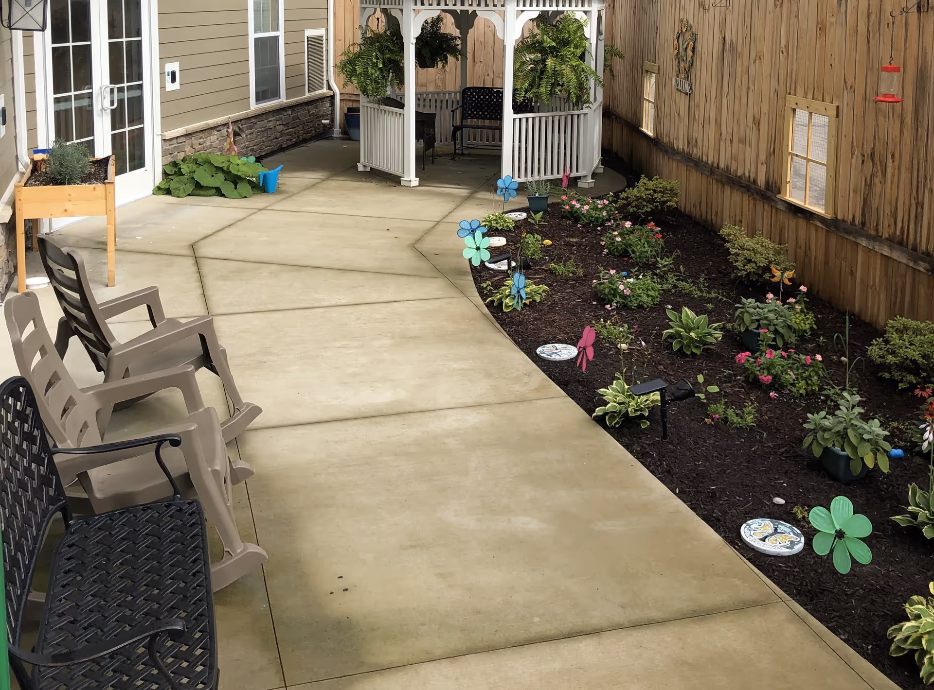 Outdoor patio area with a concrete walkway, several chairs including plastic and metal ones, a garden bed with various plants and decorative pinwheels, a white gazebo with hanging plants, and a wooden fence with small windows and a hummingbird feeder.