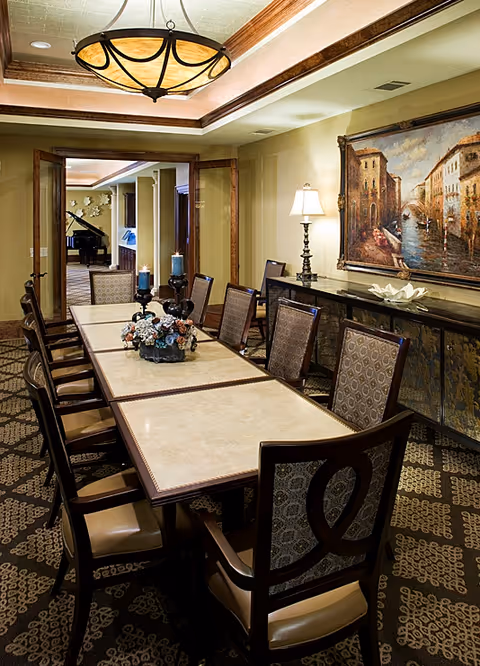 A formal dining room with a long rectangular table surrounded by upholstered chairs. The table is decorated with a floral centerpiece and candles. The room features a patterned carpet, a large framed painting on the wall, a sideboard with a lamp and decorative items, and a hanging ceiling light fixture. In the background, a grand piano is visible in an adjoining room.