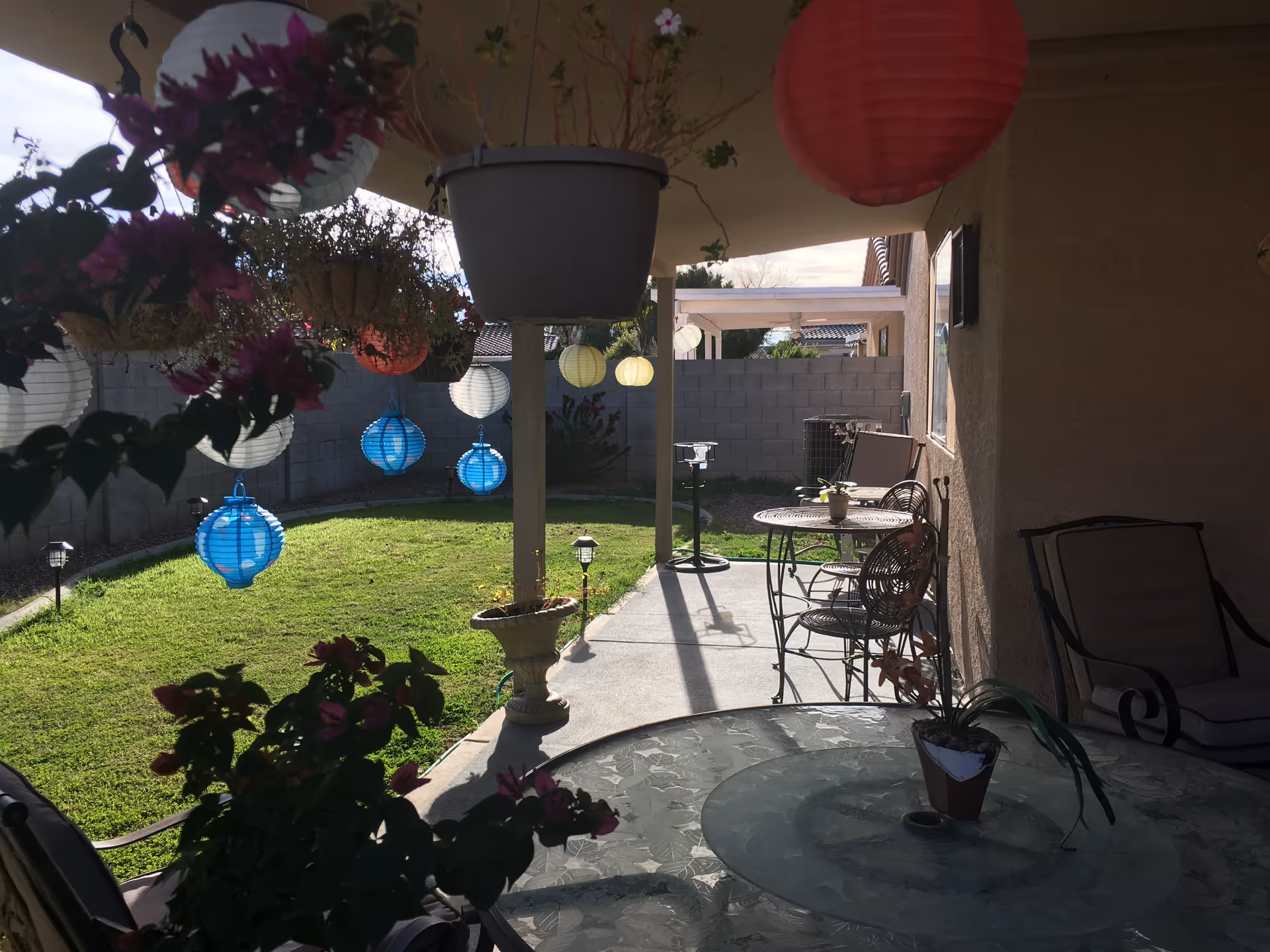 A covered patio area with hanging colorful paper lanterns and potted plants. There are metal chairs and tables on the patio, with a grassy backyard enclosed by a gray brick wall in the background.