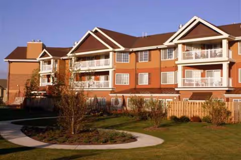Red brick senior living building with balconies and landscaped lawn, curved walkway in foreground.