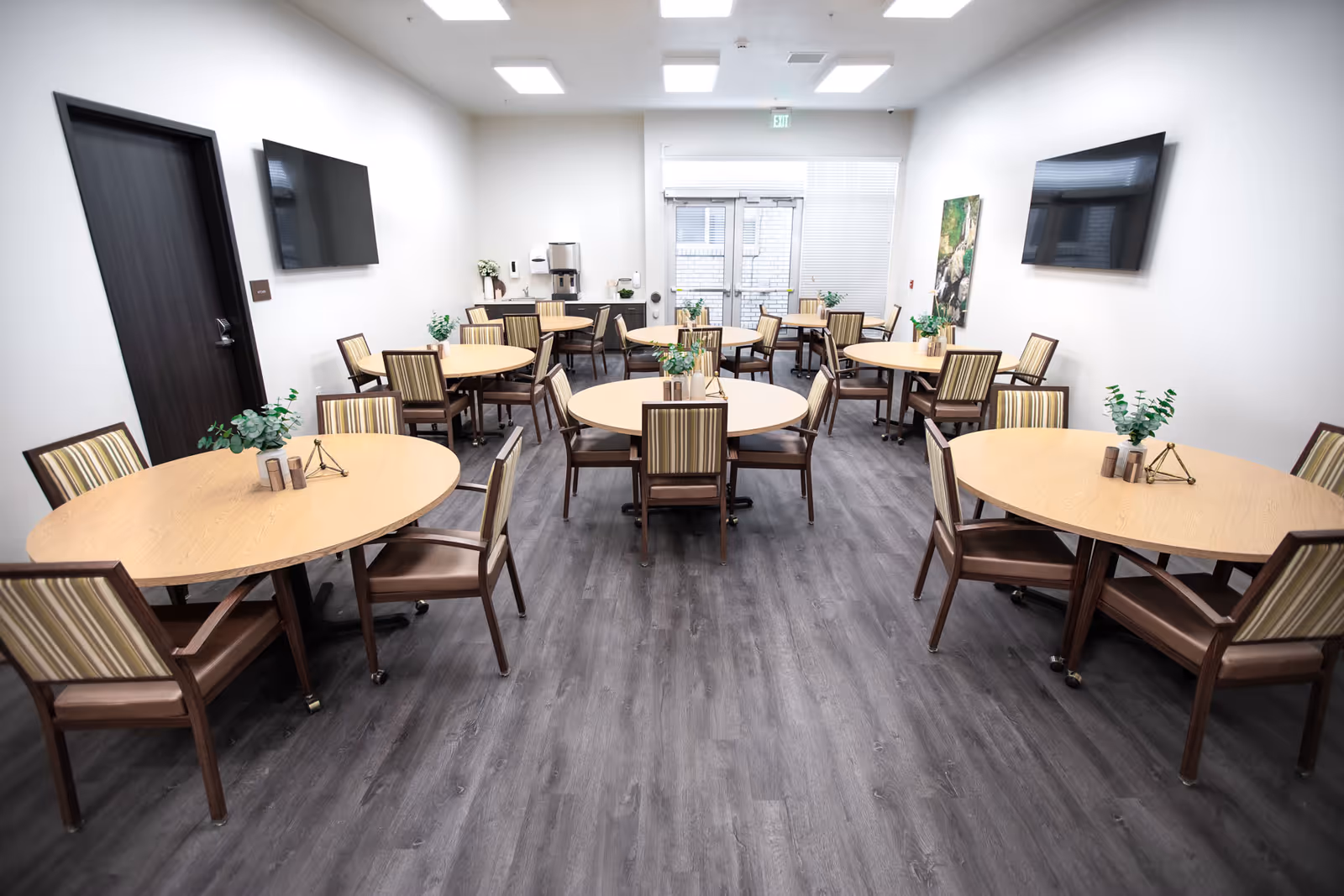 A bright dining room with multiple round wooden tables and striped cushioned chairs arranged neatly. The room has a dark wood floor, white walls, two flat-screen TVs mounted on opposite walls, and a door with windows at the far end. Small plants and decorative items are placed on the tables.