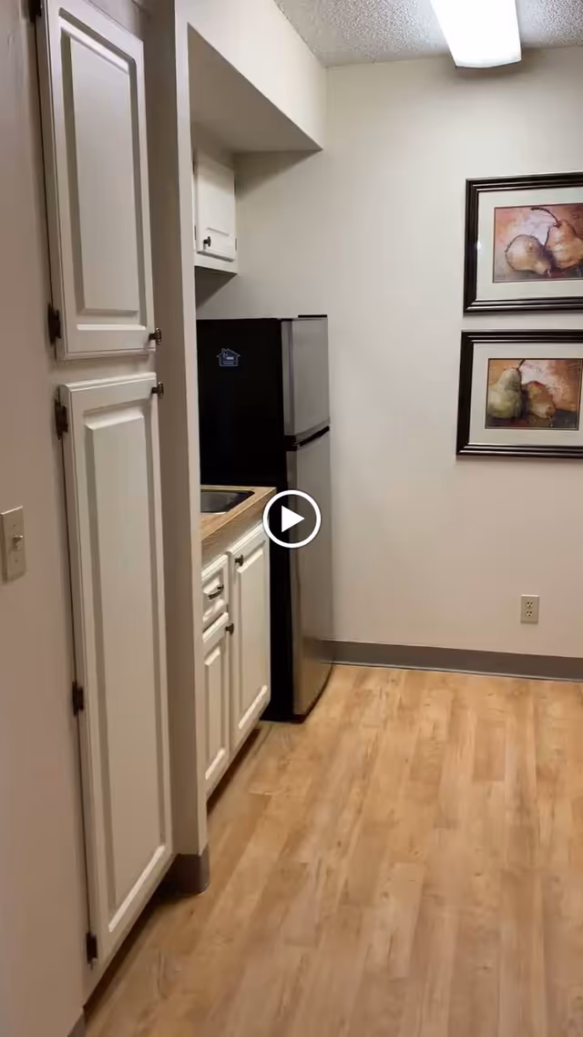 A small kitchen area with white cabinets, a stainless steel mini refrigerator, a sink, and two framed pictures of pears on the wall. The floor is light wood, and the walls are painted white.