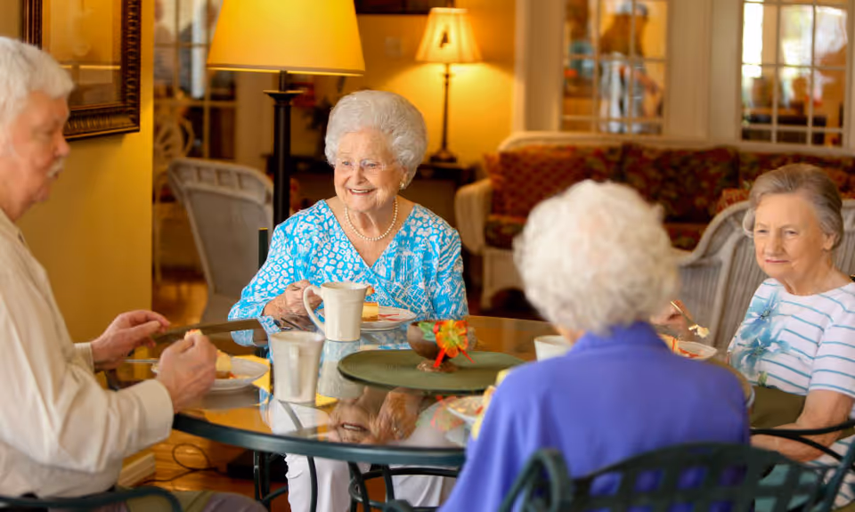 Four elderly people sitting around a glass dining table enjoying a meal together in a warmly lit room with lamps and comfortable seating in the background.