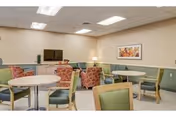 Communal seating area with tables, chairs, armchairs, a TV and framed artwork in a senior living facility.