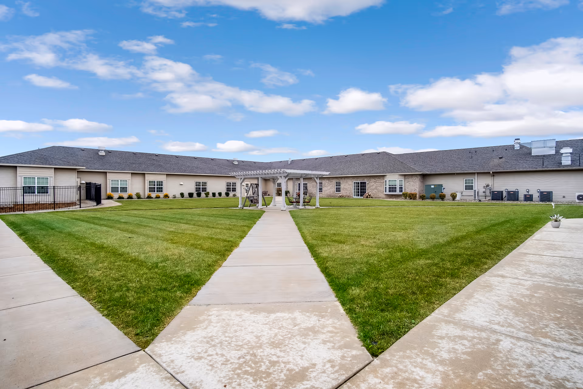 Outdoor courtyard area of Heritage Hill Assisted Living & Memory Care featuring a well-maintained green lawn, concrete walkways converging at a central pergola with a swing, surrounded by a single-story building under a partly cloudy blue sky.