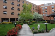 Outdoor courtyard area of a senior living facility with a concrete walkway, green grass, bushes, trees, and a white statue. The courtyard is surrounded by a multi-story brick building with many windows.