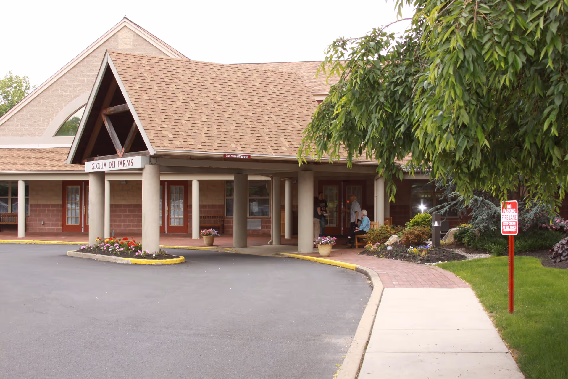 Exterior view of the entrance to Gloria Dei Farms facility with a covered drop-off area supported by columns, flower beds, a paved driveway, and a sidewalk. There are a few people near the entrance and a 'No Parking Fire Lane' sign on the right side near some greenery.