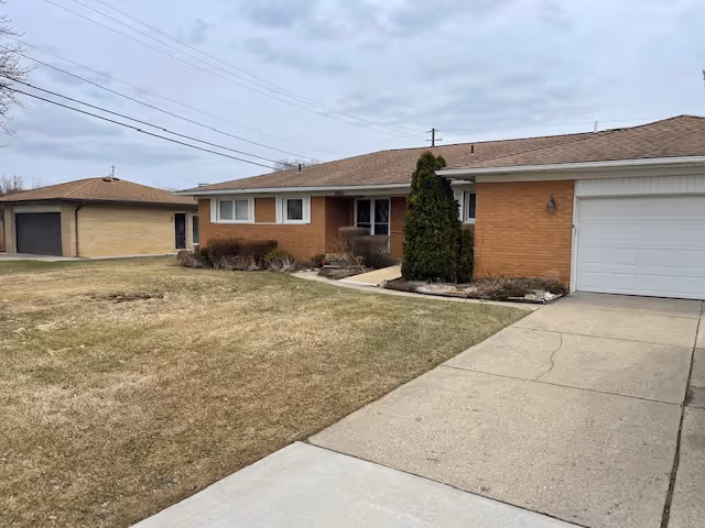 Single-story brick residential building with a garage on the right side, a concrete driveway, and a front yard with sparse grass and some bushes. The sky is cloudy.