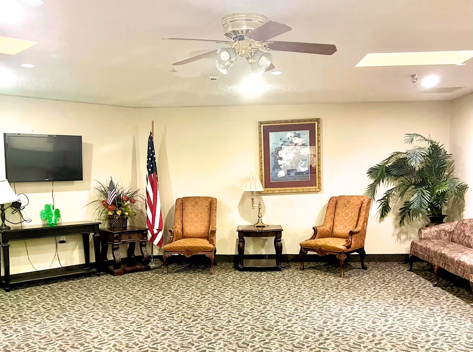 A sitting area in a nursing and rehabilitation center with two upholstered armchairs and a small table with a lamp between them. There is a framed floral picture on the wall above the table. To the left, there is a flat-screen TV mounted on the wall above a console table with a decorative plant and an American flag standing next to it. On the right side, there is a patterned sofa and a large potted plant. The room has a ceiling fan with lights and patterned carpet flooring.