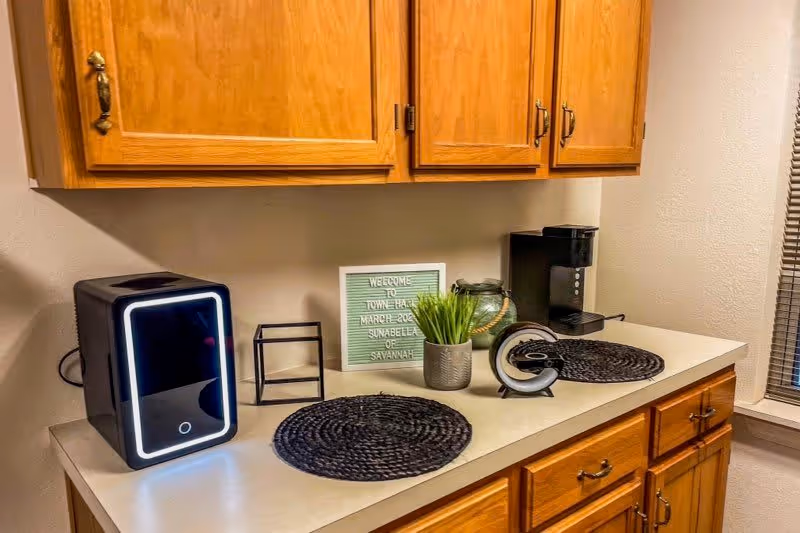 A kitchen countertop with wooden cabinets above and below. On the counter are two black woven placemats, a small black appliance with a glowing ring, a small black metal frame, a green potted plant, a decorative glass jar, a coffee maker, and a small sign that reads 'Welcome to Towne Hall March 2023 Sunabella of Savannah'. A window with blinds is visible on the right side.