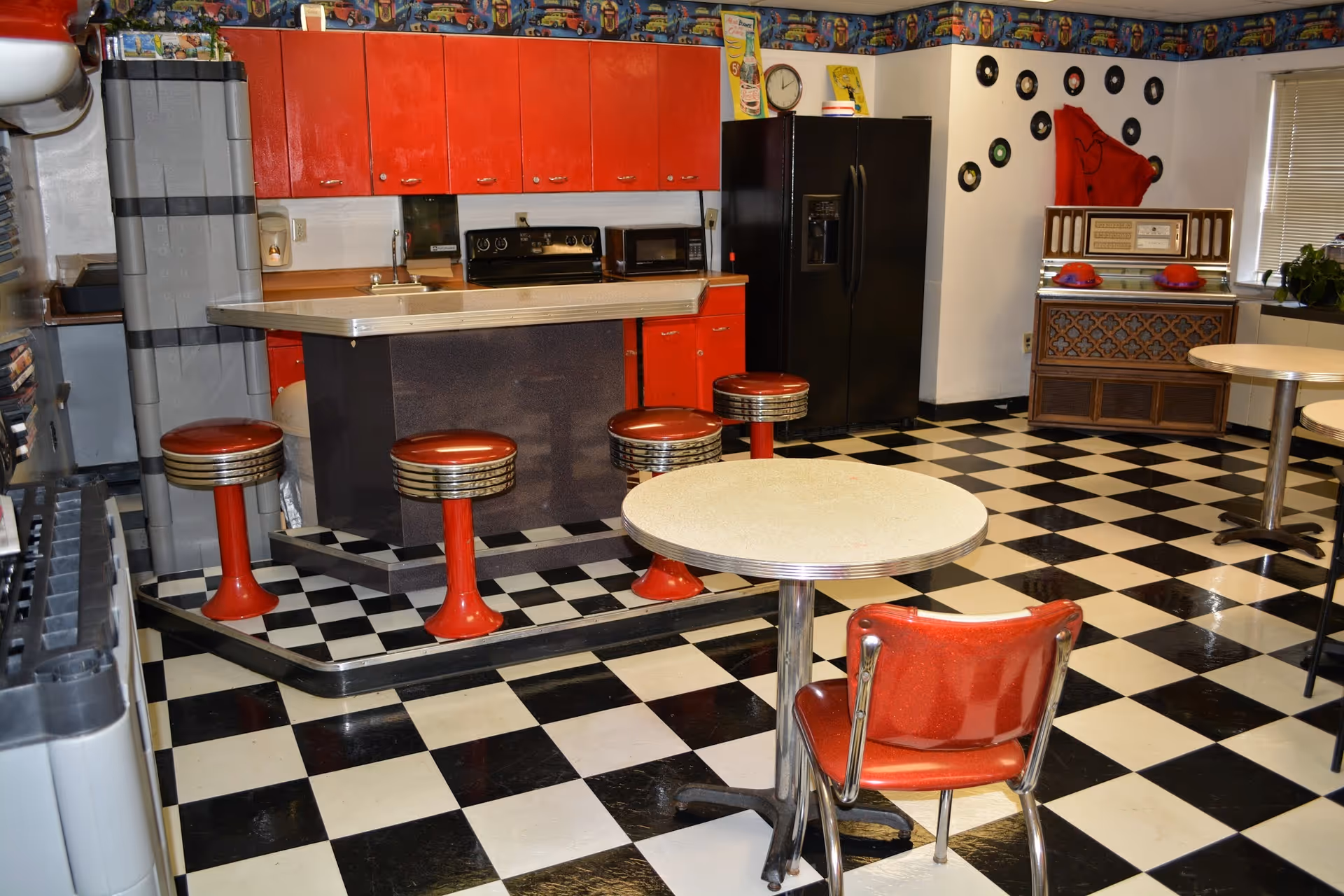 Retro-style communal dining area with a black-and-white checkered floor, red stools and chairs, a counter kitchenette and a jukebox.
