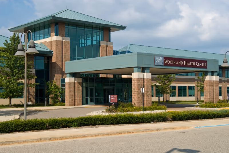 Exterior view of Woodland Health Center, a modern brick building with large glass windows and a teal roof. The entrance features a covered drop-off area with a sign reading 'Woodland Health Center'. There are trees, shrubs, and street lamps around the building under a partly cloudy sky.