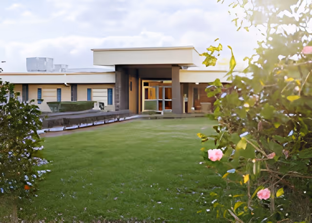 Exterior view of a single-story skilled nursing home building with a flat roof, surrounded by a green lawn and flowering bushes in the foreground under a partly cloudy sky.
