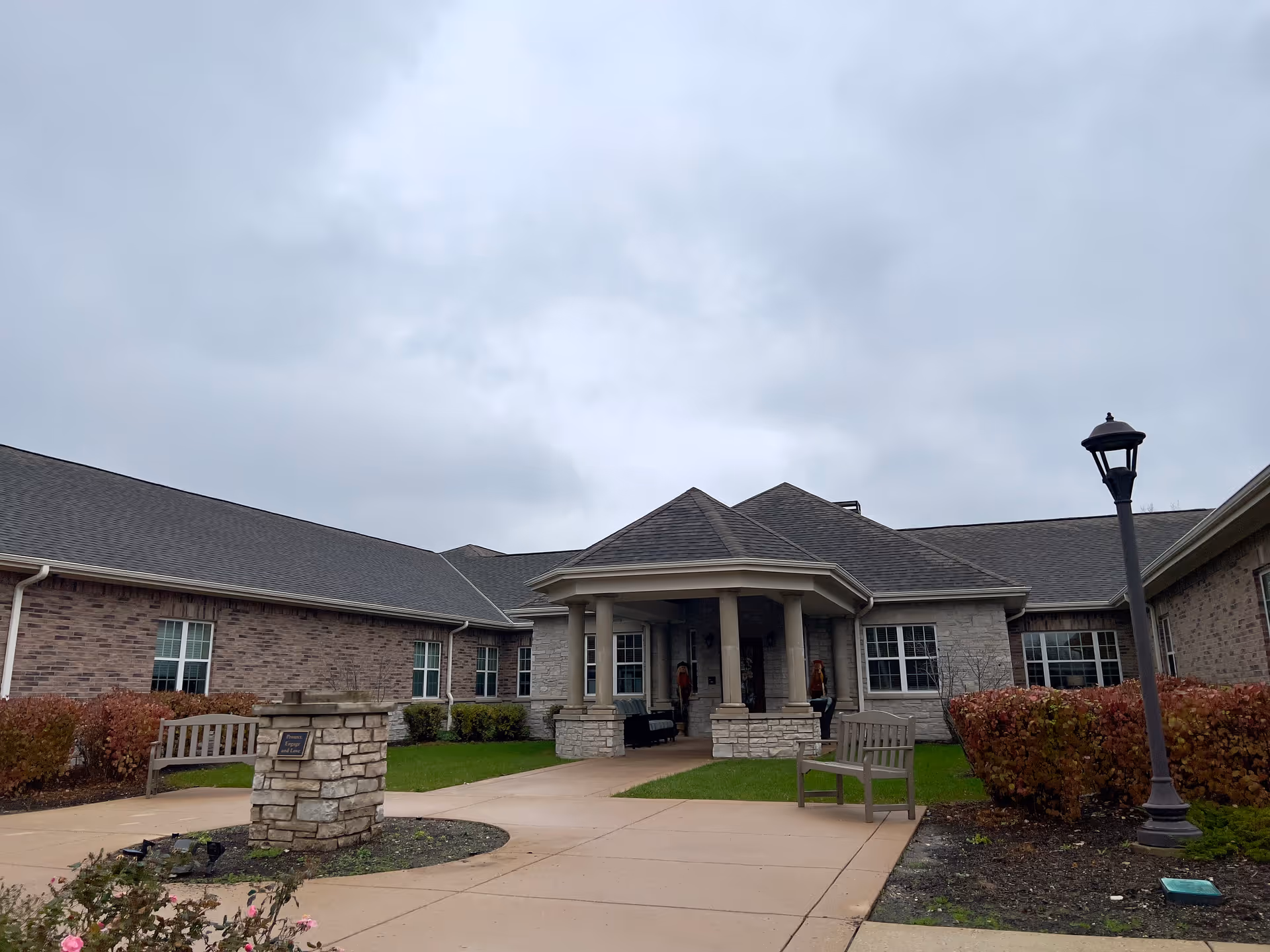 Exterior view of Franklin Place Memory Care facility showing a single-story brick building with a covered entrance supported by columns, benches on either side of the walkway, bushes, and a street lamp under a cloudy sky.