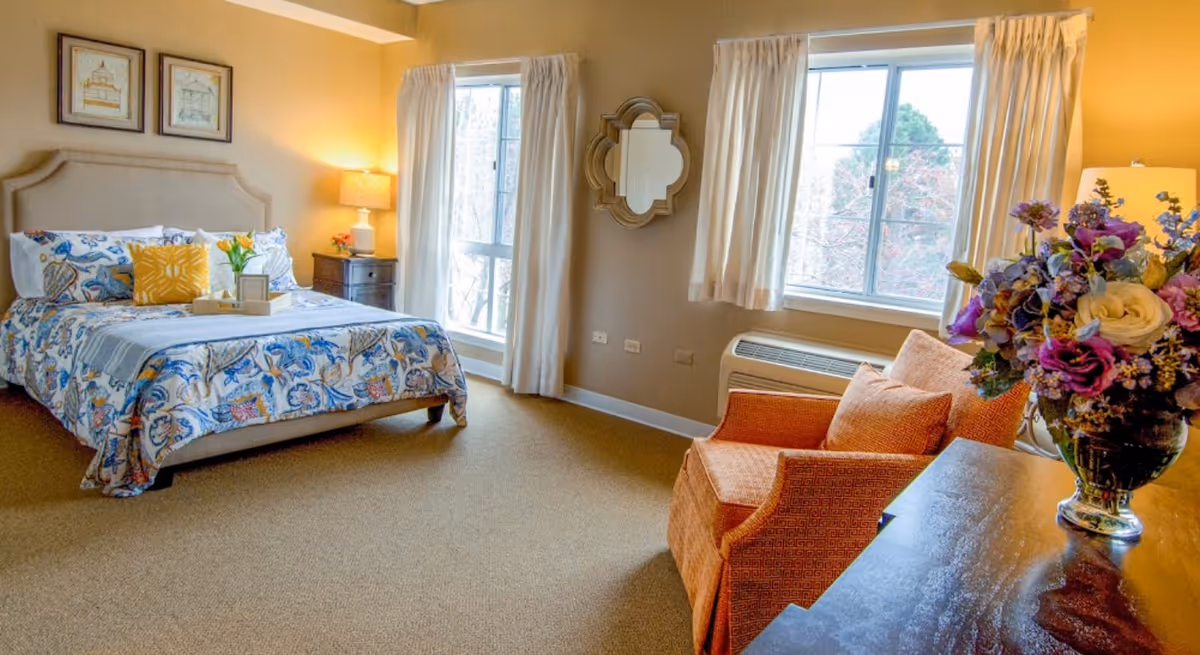 A cozy bedroom with a bed featuring a colorful floral bedspread and multiple pillows. There is a wooden nightstand with a lamp and flowers next to the bed. Two large windows with cream-colored curtains allow natural light to fill the room. An orange armchair with a matching pillow is positioned near a dark wooden dresser topped with a vase of vibrant flowers. A decorative mirror hangs on the wall between the windows.