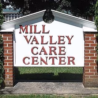 Freestanding white sign with red letters reading "MILL VALLEY CARE CENTER" set between brick posts on a lawn.