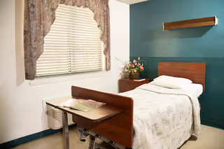 A single nursing-home bedroom with a hospital-style bed, bedside table with flowers, a window with blinds and floral curtains, and a wall shelf.