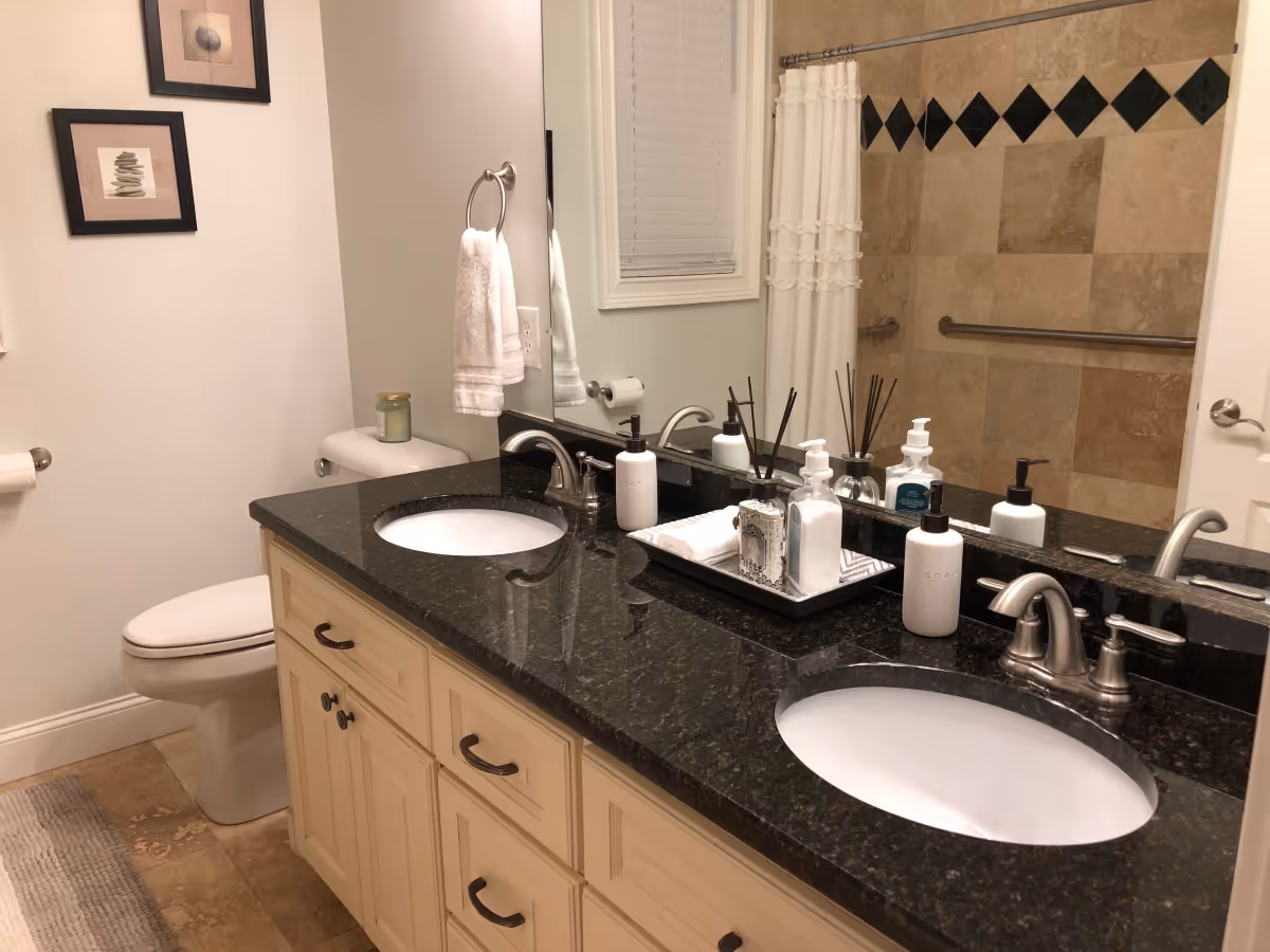 A clean and well-lit bathroom featuring a double sink vanity with black granite countertop, two silver faucets, soap dispensers, and decorative items on a tray. There is a toilet next to the vanity, a towel hanging on a ring, framed artwork on the wall, and a shower area with beige tiles and a white shower curtain.