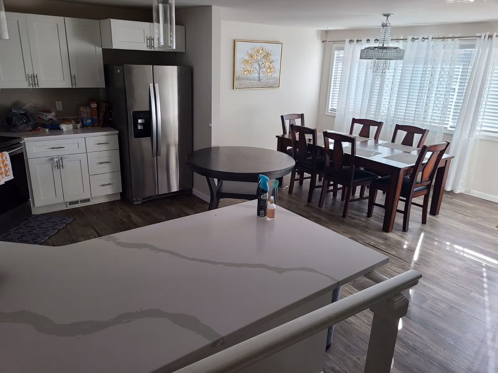 A bright kitchen and dining area with white cabinets, a stainless steel refrigerator, a round dark wood table, and a rectangular dining table with six chairs. The room has large windows with white curtains and a chandelier above the dining table. The floor is wood, and there are some cleaning supplies on the kitchen counter.