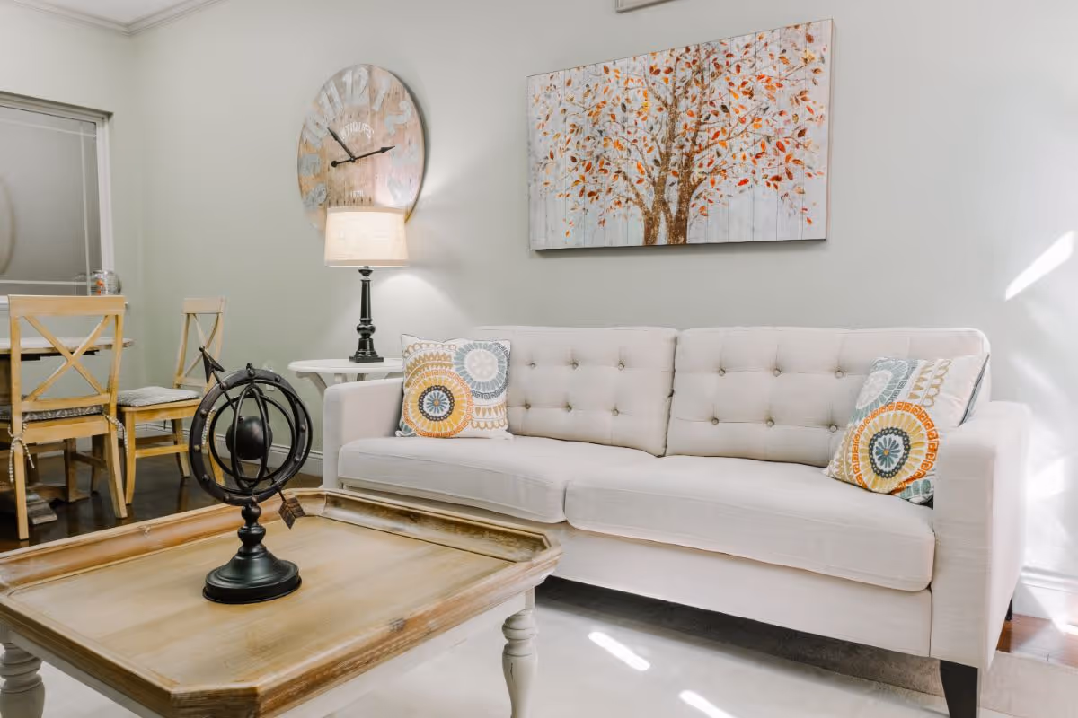 A bright living room with a beige tufted sofa adorned with two decorative pillows featuring circular patterns. A wooden coffee table with a decorative black armillary sphere sits in front of the sofa. On the wall above the sofa is a painting of a tree with orange and brown leaves. To the left, there is a dining area with a wooden table and chairs, a table lamp on a side table, and a large round wall clock.