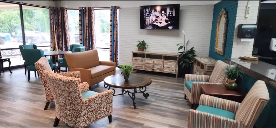 Bright senior living lounge with sofas and patterned armchairs arranged around a central coffee table, a wall-mounted TV, and large windows.