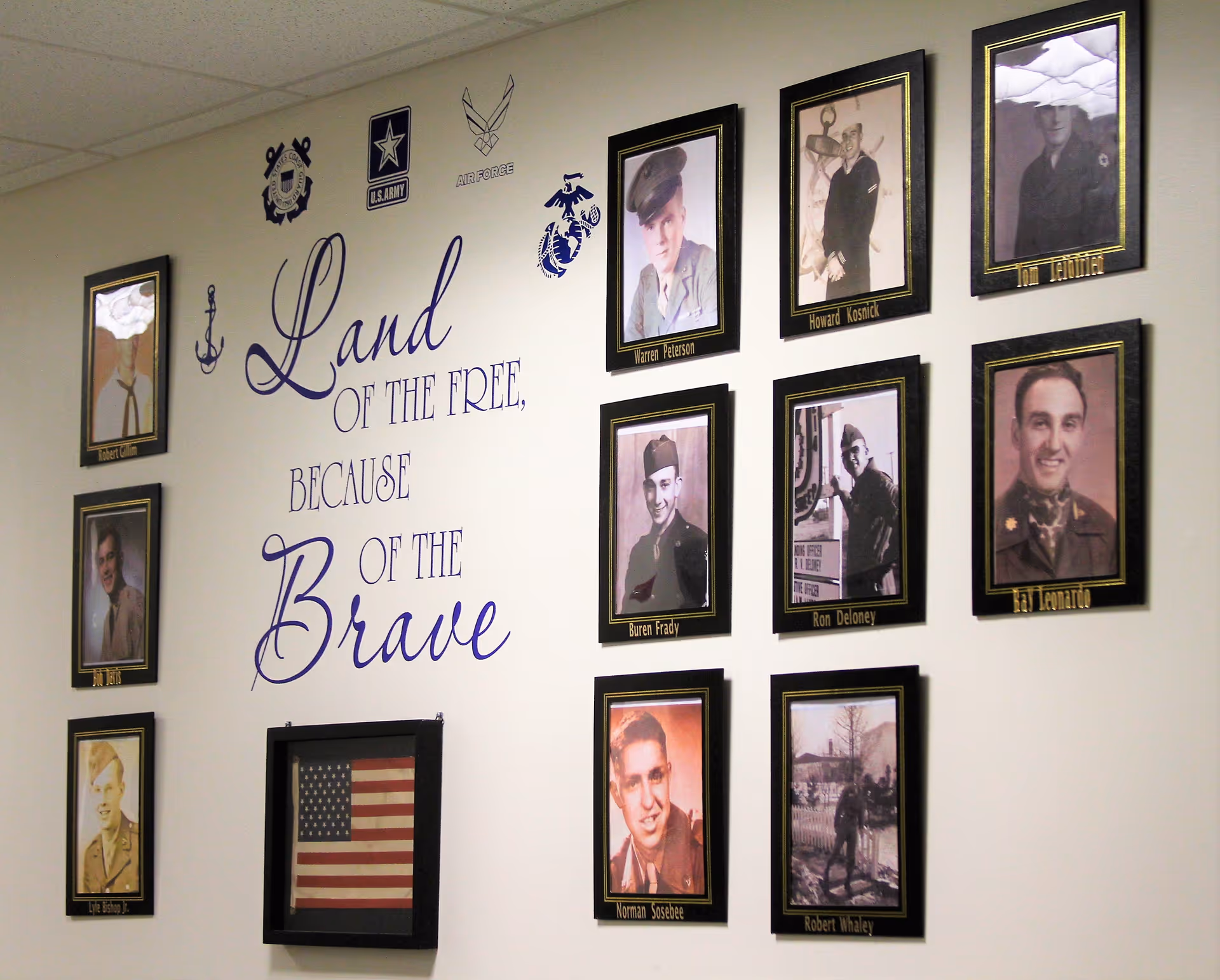 A wall display featuring framed photographs of military veterans surrounding a framed American flag. The wall has the text 'Land of the Free, Because of the Brave' along with military branch insignias including the Coast Guard, U.S. Army, Air Force, and Marine Corps.