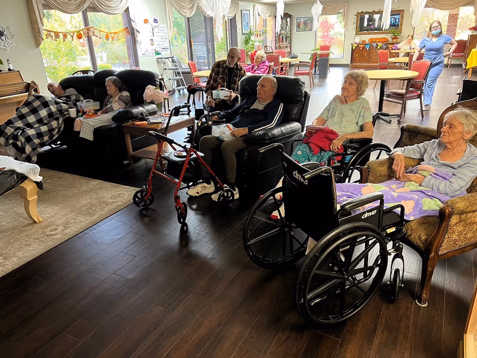 Several elderly residents seated in chairs and wheelchairs in a communal lounge area decorated for Halloween.