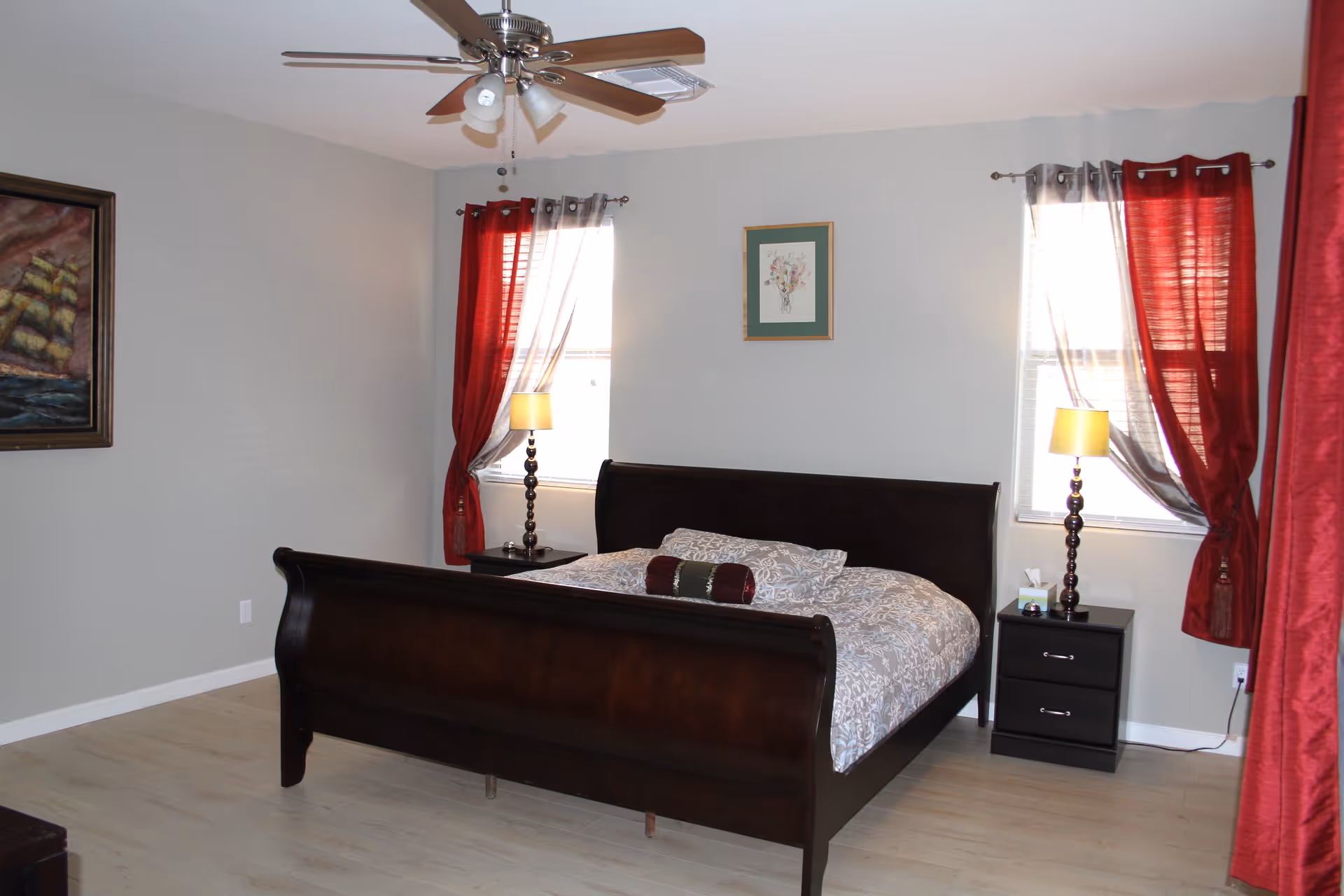 A bedroom with a dark wooden sleigh bed covered with a patterned comforter and a decorative pillow. There are two windows with red and white curtains, each with a bedside table holding a lamp. A ceiling fan with lights is mounted on the ceiling, and a framed painting of a ship is on the left wall.