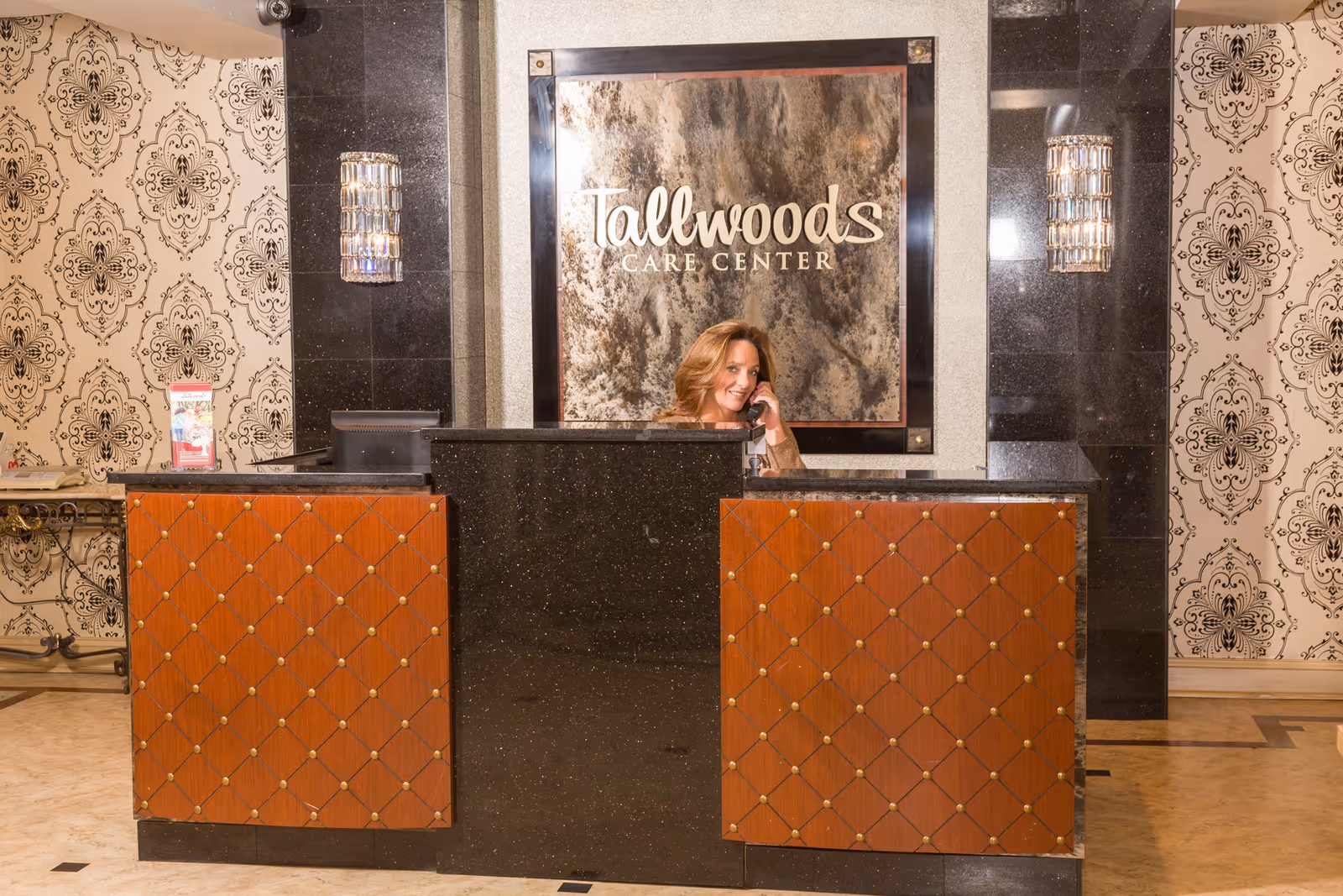 Reception area of Tallwoods Care Center featuring a woman speaking on the phone behind a black and wood-paneled front desk. The background has a large sign with the facility's name and decorative wallpaper with a black and white pattern. Two wall-mounted light fixtures flank the sign.