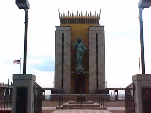 Statue stands in front of a tall brick memorial with a spiked crown, flanked by flagpoles.