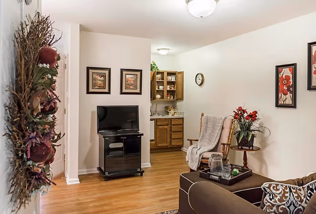 Cozy living room with a TV on a stand, an armchair and ottoman, decorative wall art and a small kitchenette visible in the background.