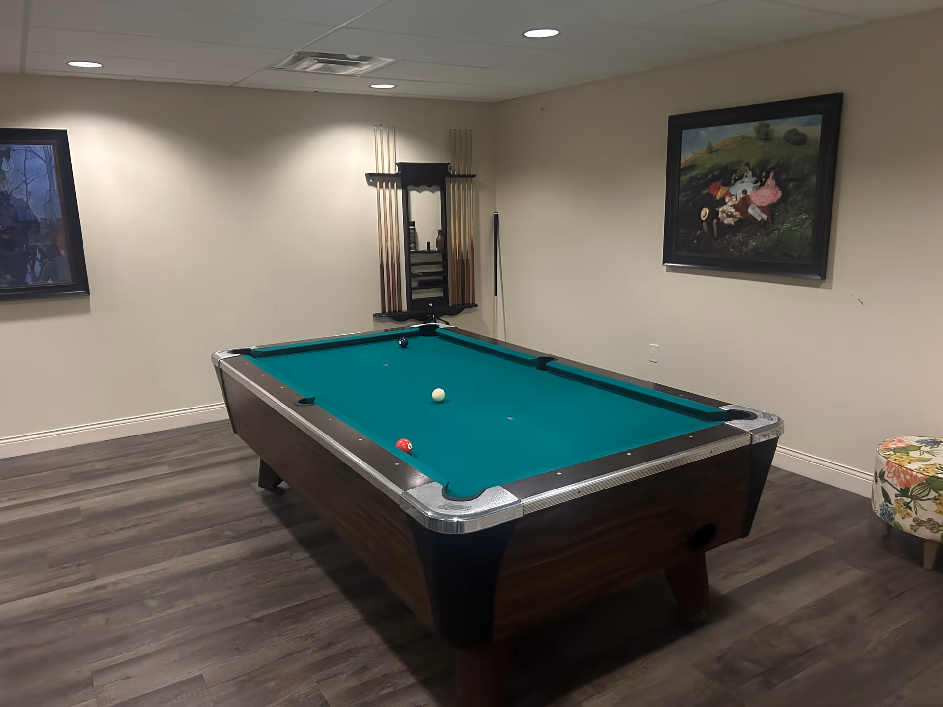 A room with a pool table in the center, featuring a green felt surface and a few billiard balls on it. The walls are beige with two framed paintings hanging on them. A rack with pool cues and accessories is mounted on the wall behind the table. The floor has wood-like vinyl or laminate flooring, and there is a floral-patterned round ottoman in the corner.