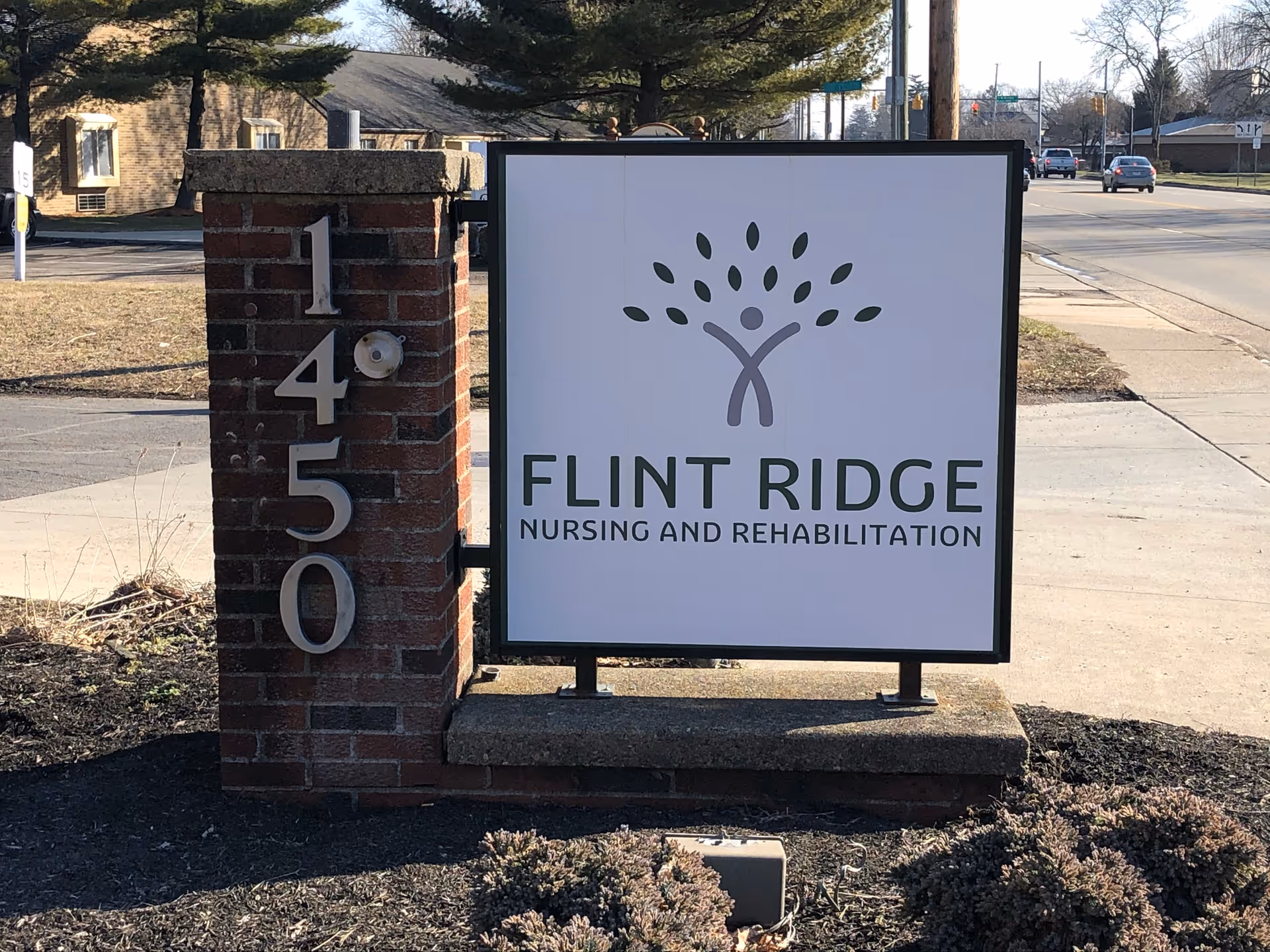 Outdoor sign for Flint Ridge Nursing and Rehabilitation mounted on a brick pillar with the address number 1450 displayed vertically. The sign features a logo of a stylized person with leaves above them, resembling a tree. The background shows a sidewalk, street, and some buildings in the distance.