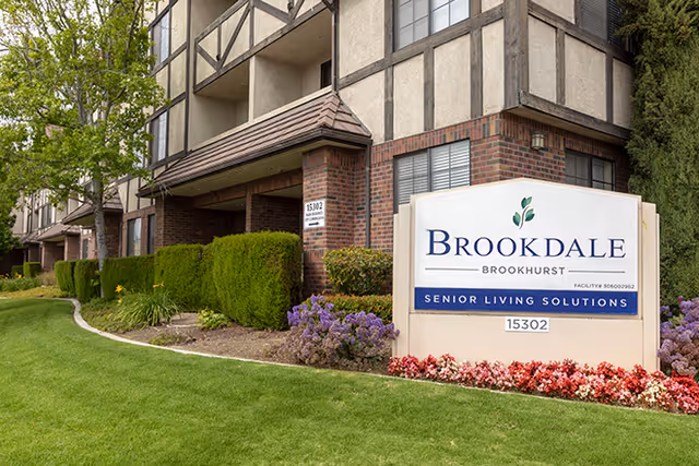 Exterior view of the Brookdale Brookhurst senior living facility showing a large sign with the facility name and address, surrounded by well-maintained landscaping including green grass, bushes, and flowers. The building has a brick and timber facade with balconies and windows.