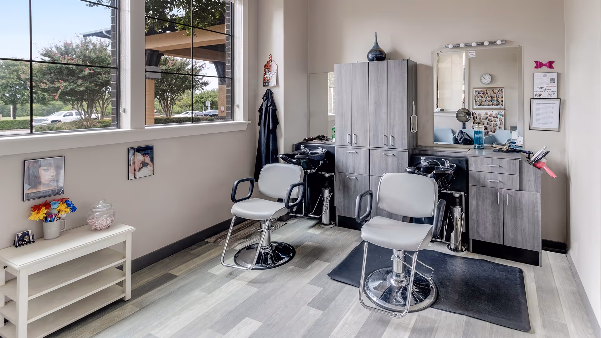 Interior view of a hair salon area in a senior living facility with two salon chairs, a large mirror, and cabinetry. There are hair washing sinks behind the chairs, a window showing trees and parked cars outside, and a small white table with colorful flowers and framed pictures on the left side.