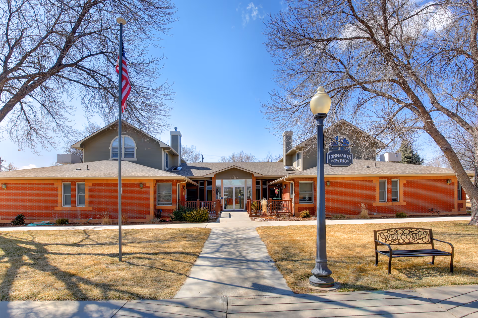 Front exterior view of Cinnamon Park facility with a brick building, a pathway leading to the entrance, an American flag on a flagpole, a street lamp, and a bench with the word 'Welcome' on it. Leafless trees are visible around the building under a clear blue sky.