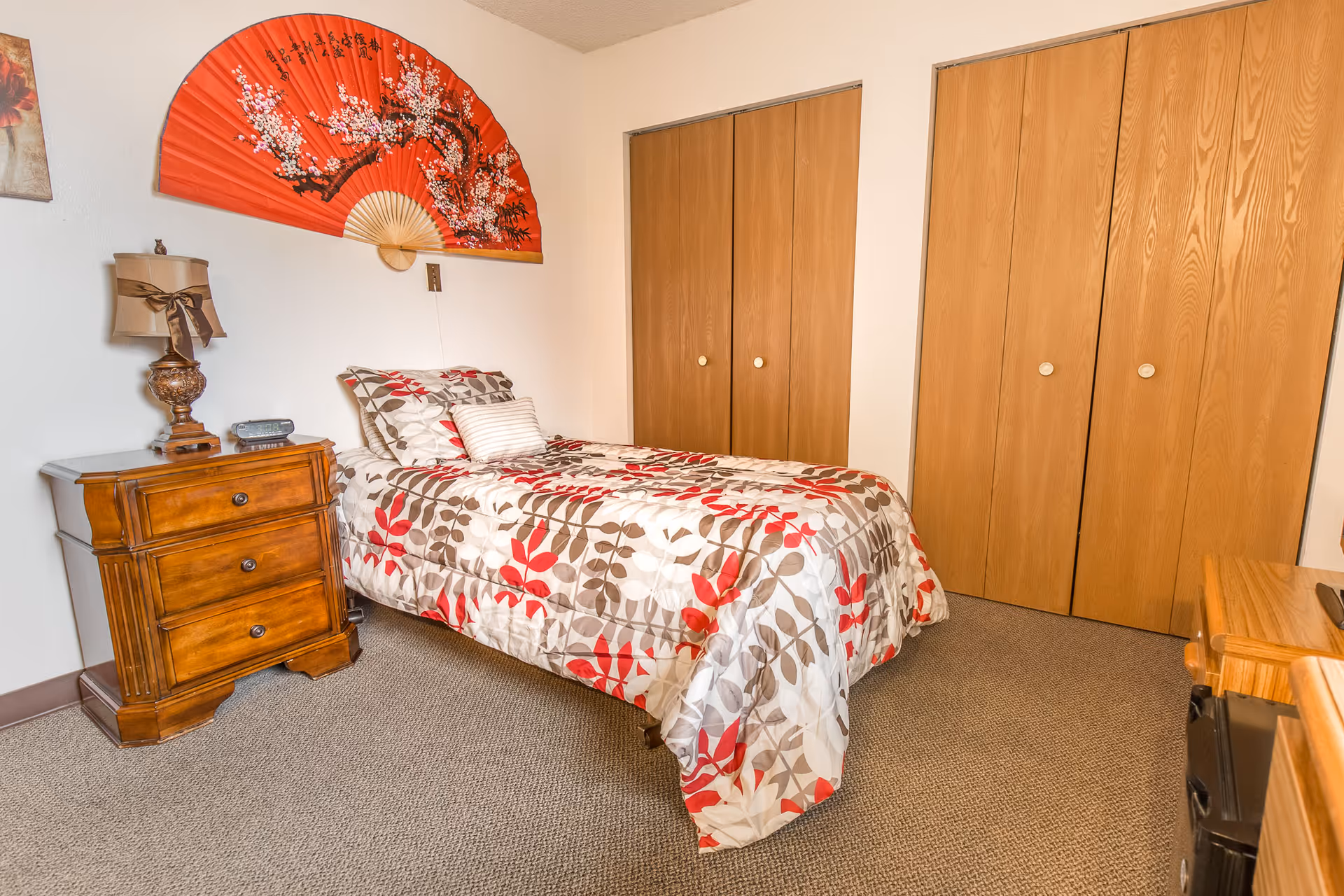 A bedroom with a single bed covered in a floral patterned bedspread in shades of red, white, and brown. There is a wooden nightstand with a decorative lamp and a clock next to the bed. Above the bed, a large red decorative fan with floral designs is mounted on the wall. The room has beige carpet and two wooden closets with sliding doors.