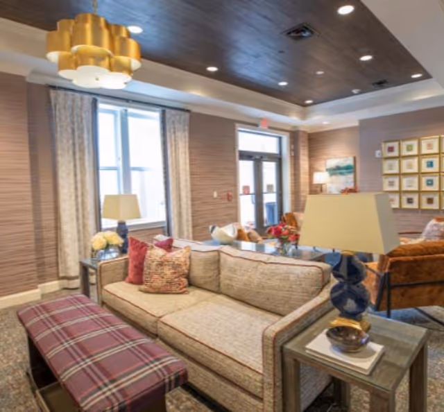 A cozy living room area with a beige sofa adorned with patterned pillows, a plaid upholstered bench, and a side table with a lamp and decorative items. The room features large windows with curtains, a wooden ceiling with recessed lighting, and a modern chandelier. There are additional chairs and artwork on the walls, creating a warm and inviting atmosphere.
