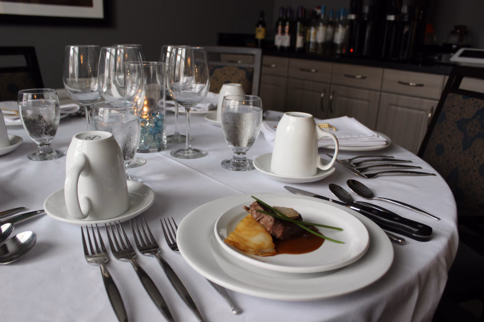 A round dining table set with white tablecloth, multiple forks, knives, spoons, upside-down white coffee cups on saucers, water glasses, wine glasses, and a plate with a small serving of meat, sauce, and garnish. In the background, there is a cabinet with bottles and other dining items.